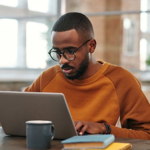 Young man with glasses works on laptop at a wooden table indoors. Warm, productive atmosphere.