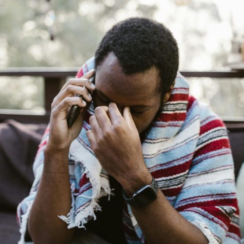 A man wrapped in a blanket looks distressed while talking on the phone outdoors.
