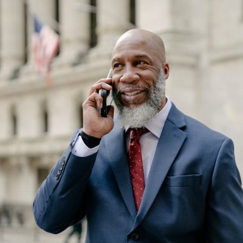 A mature businessman in a blue suit smiling during a phone call outside an iconic building.