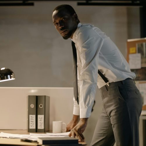 A detective stands at a desk, analyzing documents, in a well-lit office environment.